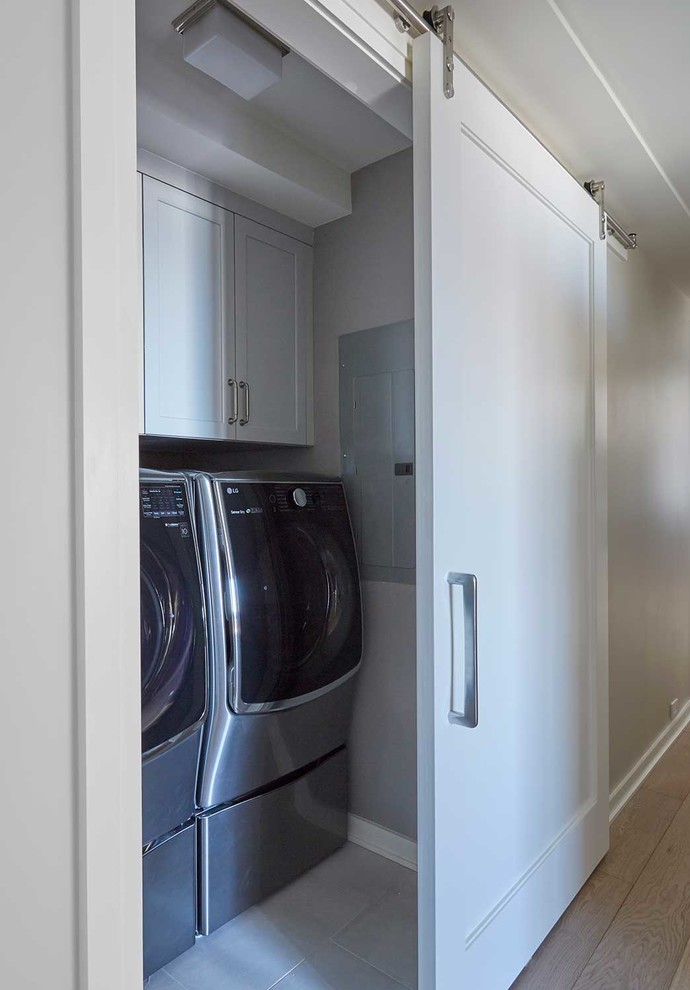 Modelo de armario lavadero lineal actual pequeño con armarios estilo shaker, puertas de armario blancas, paredes grises, suelo de baldosas de porcelana, lavadora y secadora juntas y suelo beige