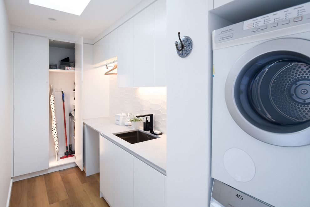 シドニーにある高級な広いモダンスタイルのおしゃれな洗濯室 (L型、アンダーカウンターシンク、フラットパネル扉のキャビネット、白いキャビネット、クオーツストーンカウンター、白いキッチンパネル、セラミックタイルのキッチンパネル、白い壁、無垢フローリング、上下配置の洗濯機・乾燥機、茶色い床、グレーのキッチンカウンター) の写真