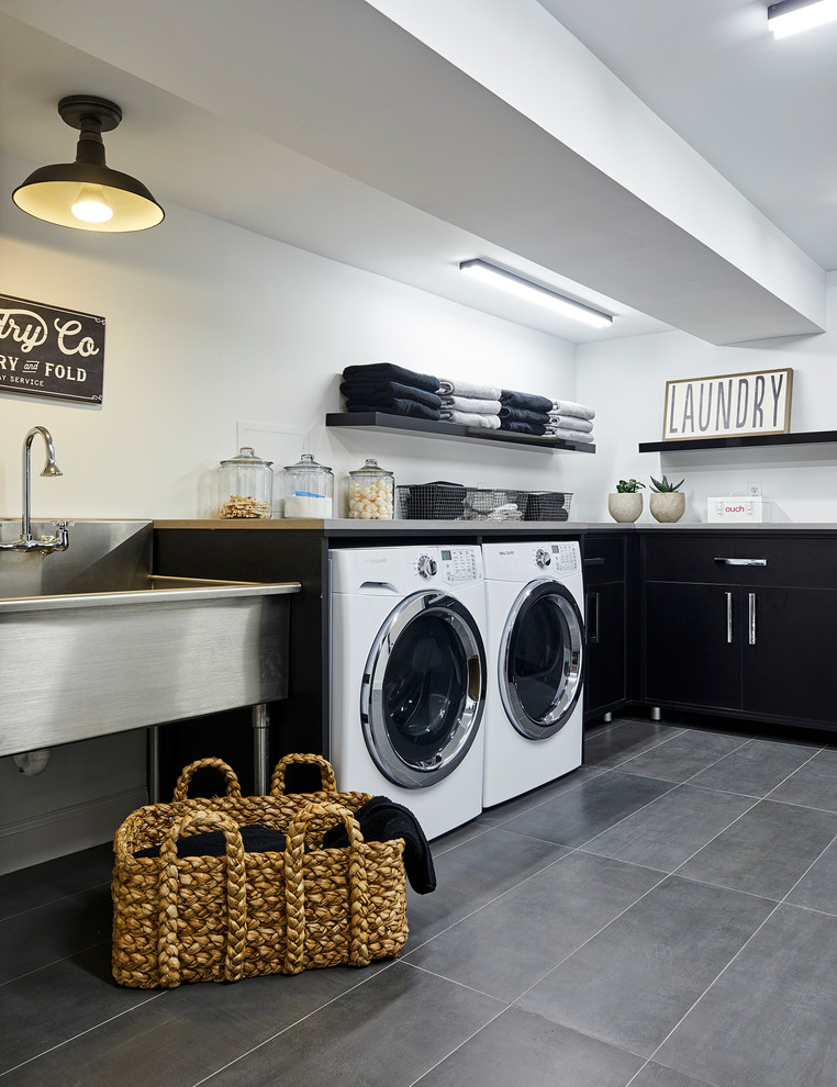 ニューヨークにある広いコンテンポラリースタイルのおしゃれな洗濯室 (L型、スロップシンク、フラットパネル扉のキャビネット、黒いキャビネット、白い壁、磁器タイルの床、左右配置の洗濯機・乾燥機、グレーの床) の写真