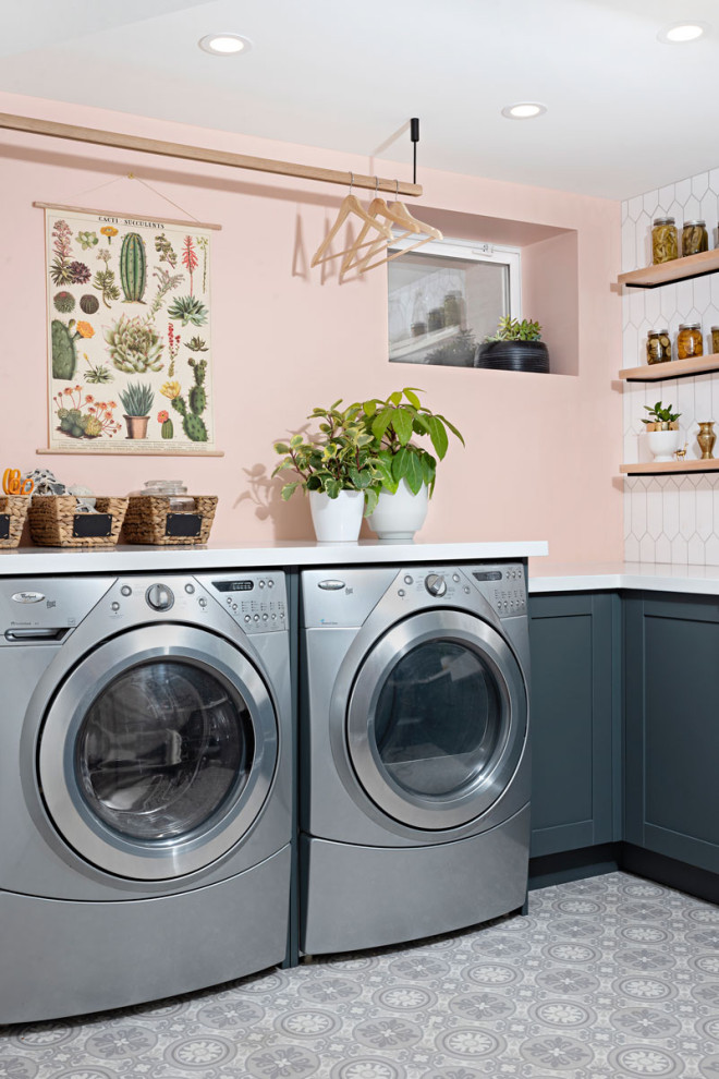 Lantlig inredning av en vita l-formad vitt tvättstuga, med skåp i shakerstil, blå skåp, bänkskiva i kvarts, linoleumgolv, en tvättmaskin och torktumlare bredvid varandra, grått golv och rosa väggar