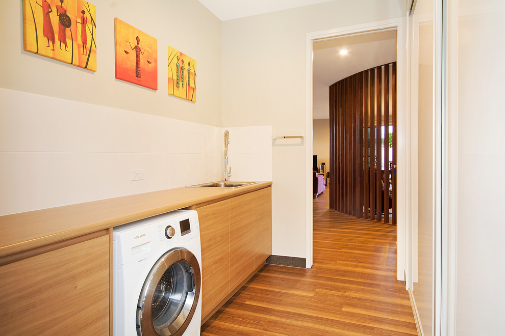 Foto på en funkis linjär tvättstuga, med en nedsänkt diskho, laminatbänkskiva, vinylgolv, släta luckor, en tvättmaskin och torktumlare bredvid varandra, skåp i mellenmörkt trä och beige väggar