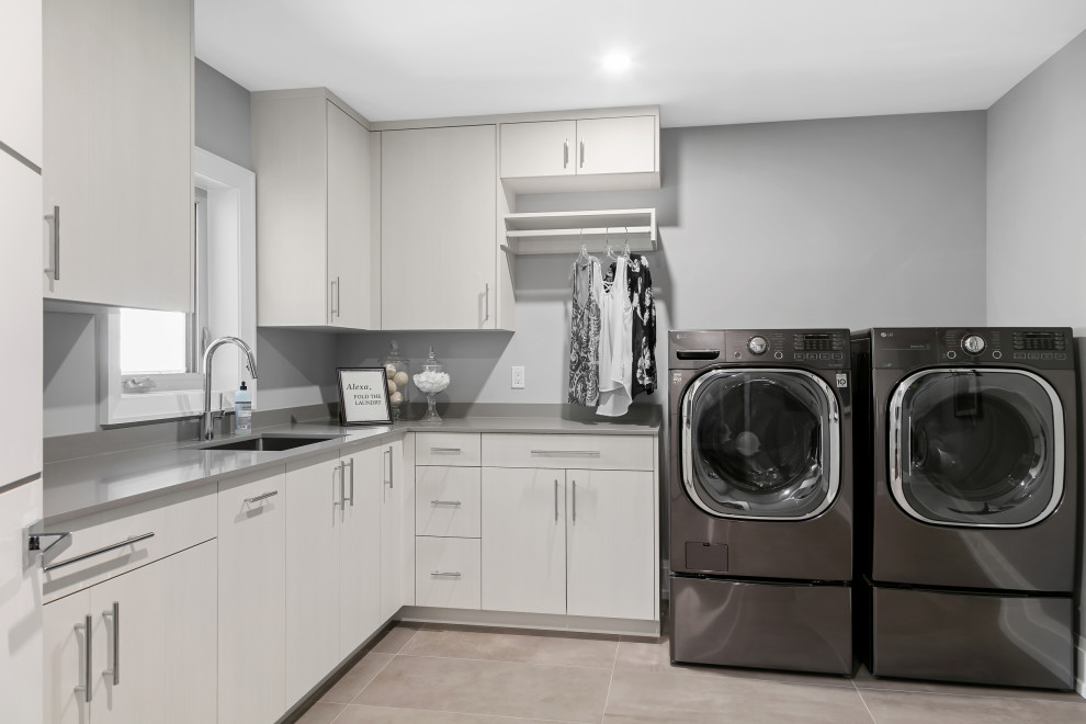 Idéer för en stor modern grå l-formad tvättstuga enbart för tvätt, med en undermonterad diskho, släta luckor, vita skåp, bänkskiva i kvarts, grått stänkskydd, grå väggar, klinkergolv i porslin, en tvättmaskin och torktumlare bredvid varandra och grått golv