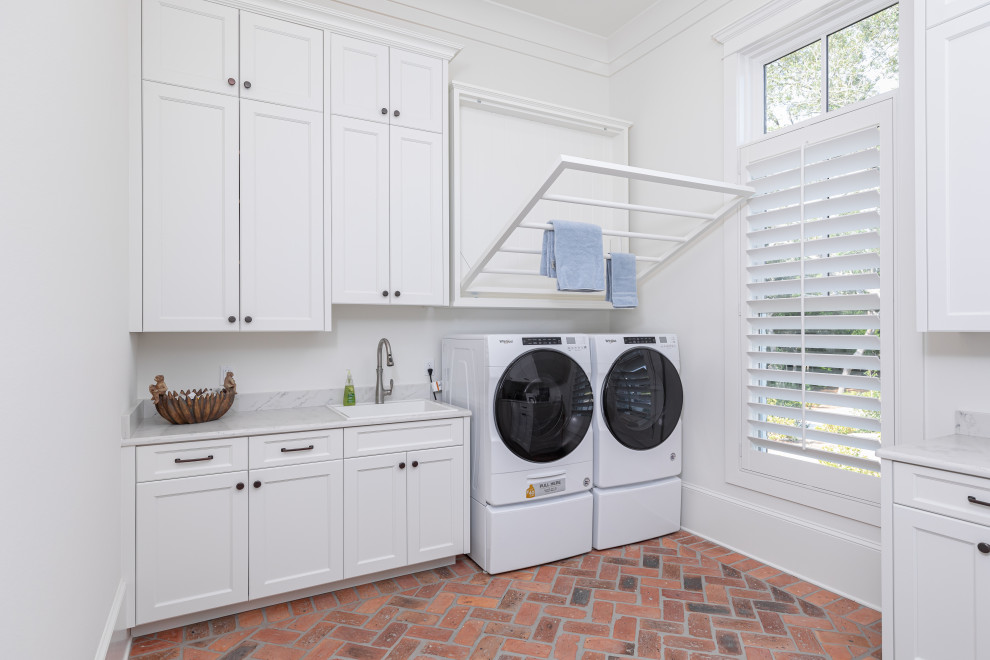 Modern inredning av en stor vita l-formad vitt tvättstuga enbart för tvätt, med en nedsänkt diskho, luckor med infälld panel, vita skåp, marmorbänkskiva, vita väggar, tegelgolv, en tvättmaskin och torktumlare bredvid varandra och brunt golv