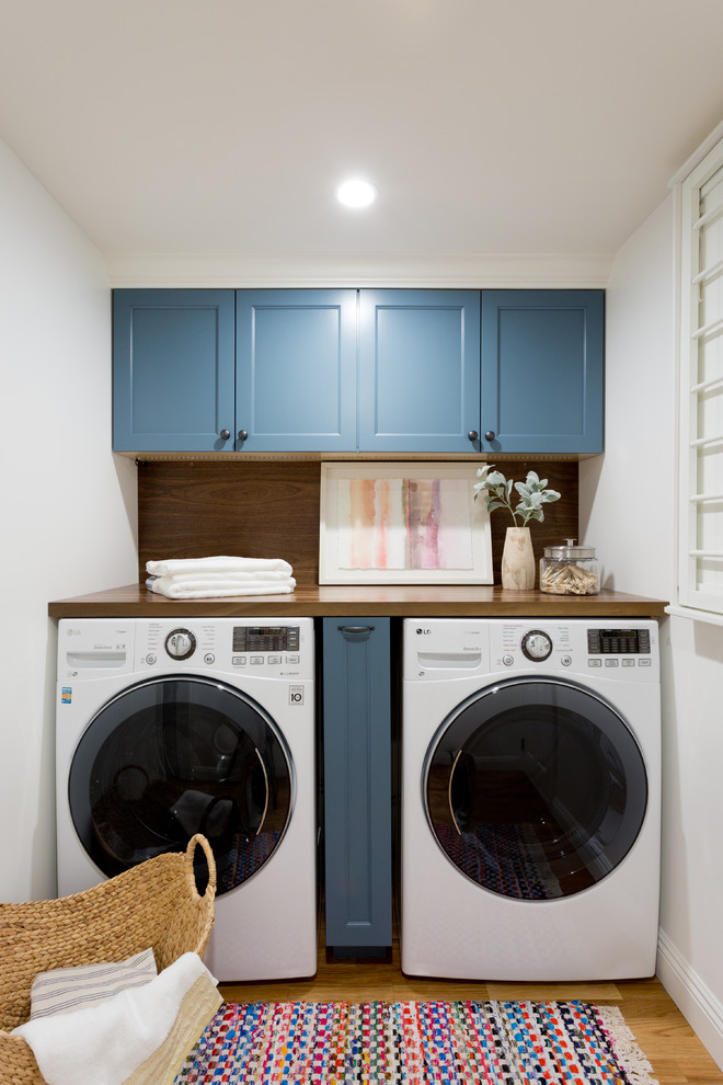 Modern inredning av en tvättstuga enbart för tvätt, med blå skåp, träbänkskiva, mellanmörkt trägolv och en tvättmaskin och torktumlare bredvid varandra