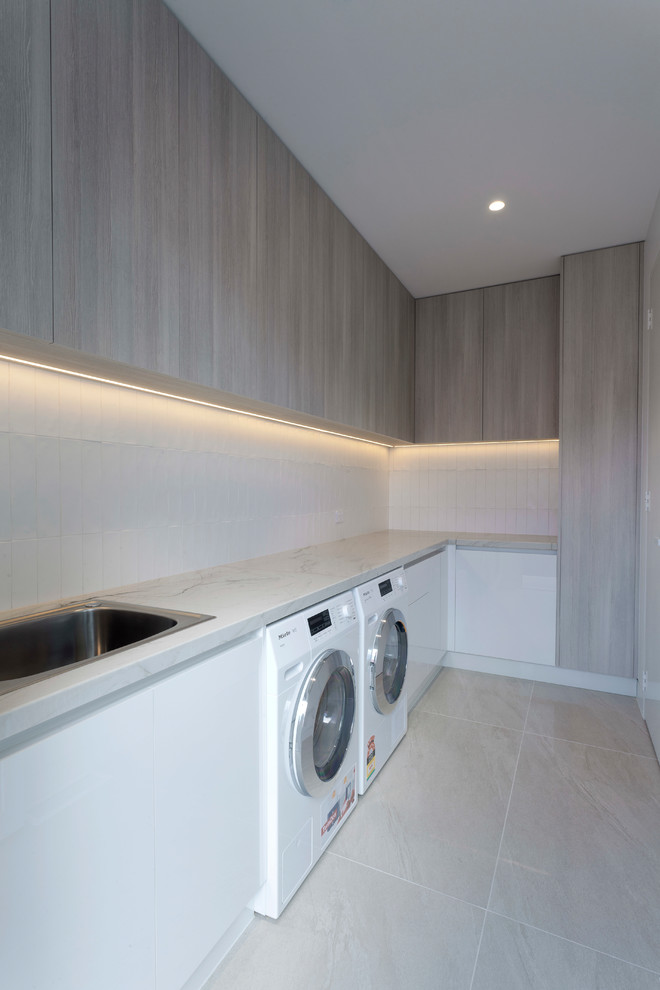 Inspiration for a large modern l-shaped laundry room in Melbourne with a drop-in sink, flat-panel cabinets, distressed cabinets, quartzite benchtops, ceramic splashback, porcelain floors, a side-by-side washer and dryer and beige floor.