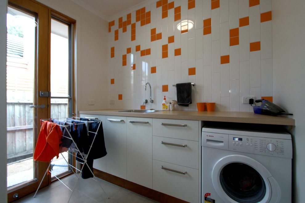 Modelo de cuarto de lavado lineal moderno pequeño con fregadero encastrado, armarios con paneles lisos, puertas de armario blancas, encimera de madera y lavadora y secadora juntas