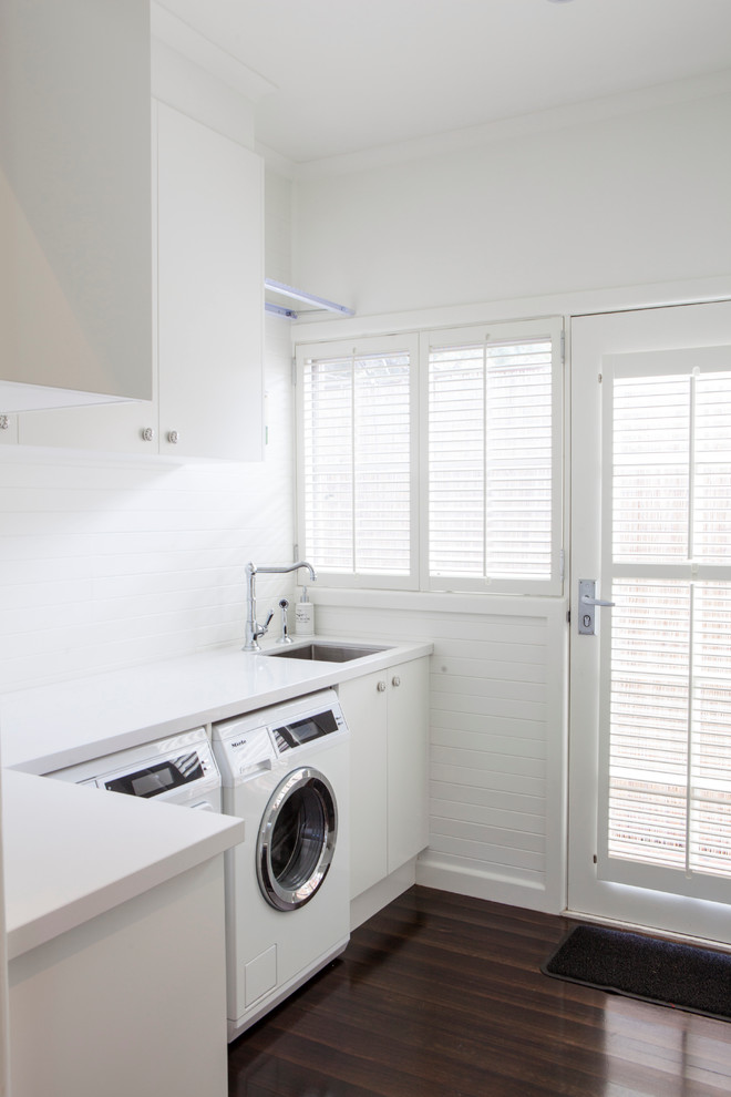 Moderner Hauswirtschaftsraum mit weißen Schränken, Waschmaschine und Trockner nebeneinander, dunklem Holzboden und weißer Arbeitsplatte in Melbourne