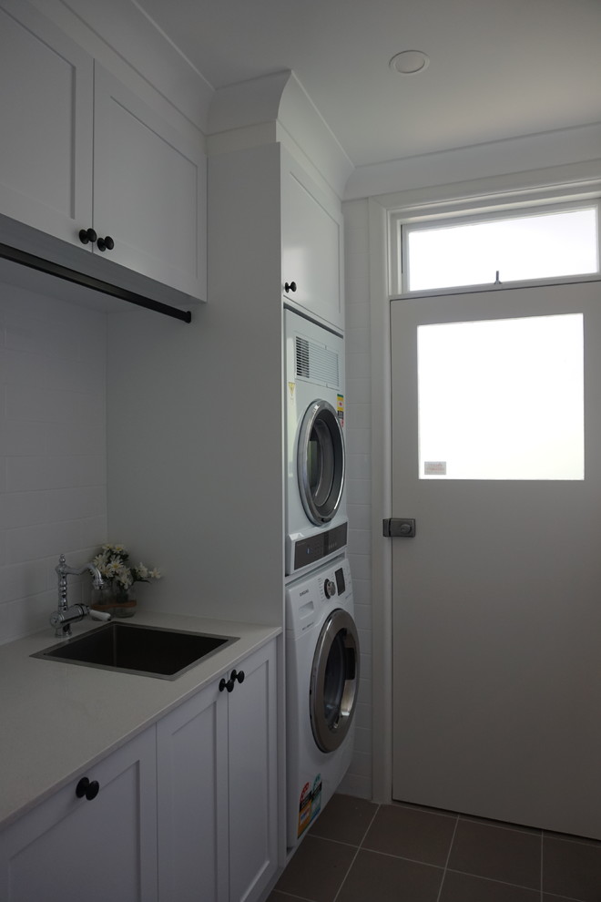 Einzeilige, Kleine Moderne Waschküche mit Waschbecken, Schrankfronten mit vertiefter Füllung, weißen Schränken, Quarzwerkstein-Arbeitsplatte, weißer Wandfarbe, Porzellan-Bodenfliesen, Waschmaschine und Trockner gestapelt und grauem Boden in Sydney