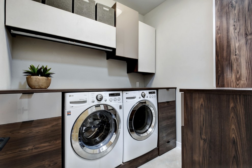 Modern inredning av en mellanstor bruna brunt tvättstuga enbart för tvätt, med släta luckor, skåp i mörkt trä, träbänkskiva, grå väggar, betonggolv, en tvättmaskin och torktumlare bredvid varandra och grått golv