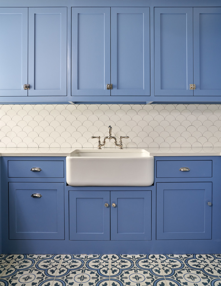 Inspiration for a mid-sized transitional l-shaped blue floor and ceramic tile dedicated laundry room remodel in Dallas with a farmhouse sink, shaker cabinets, blue cabinets, white walls, white countertops and solid surface countertops