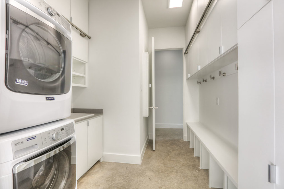 Photo of a medium sized modern galley utility room in Houston with a built-in sink, flat-panel cabinets, white cabinets, white walls, concrete flooring, a stacked washer and dryer, beige floors and laminate countertops.