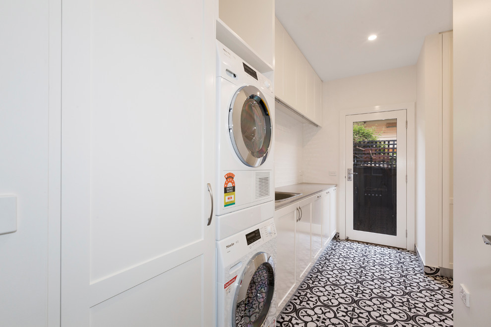 Example of a small trendy single-wall porcelain tile dedicated laundry room design in Melbourne with a single-bowl sink, white cabinets, granite countertops, white walls, a stacked washer/dryer and recessed-panel cabinets