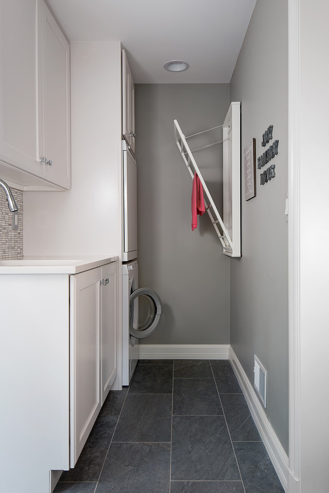 Dedicated laundry room - mid-sized transitional single-wall ceramic tile and gray floor dedicated laundry room idea in Detroit with white cabinets, gray walls, a stacked washer/dryer, recessed-panel cabinets, an undermount sink, solid surface countertops and white countertops