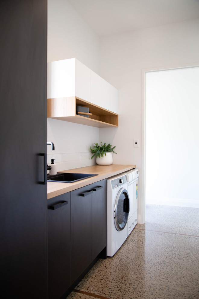 Immagine di una lavanderia multiuso minimalista di medie dimensioni con lavello da incasso, ante lisce, ante nere, top in laminato, pareti bianche, pavimento in cemento e lavatrice e asciugatrice affiancate