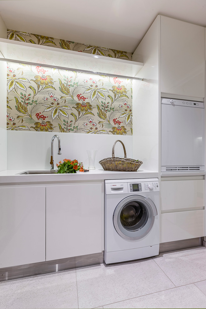 Example of a mid-sized trendy galley porcelain tile dedicated laundry room design in Auckland with a drop-in sink, flat-panel cabinets, white cabinets, quartz countertops, multicolored walls and a stacked washer/dryer