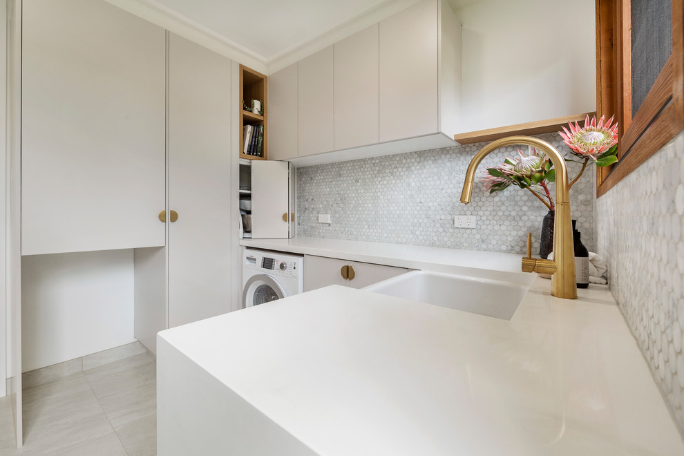 This is an example of a medium sized contemporary u-shaped utility room in Melbourne with a belfast sink, flat-panel cabinets, white cabinets, engineered stone countertops, white walls, porcelain flooring, a side by side washer and dryer, grey floors and white worktops.