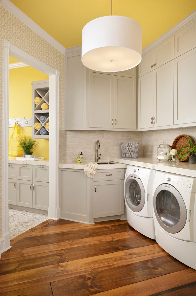 Kleine Klassische Waschküche in L-Form mit Schrankfronten im Shaker-Stil, grauen Schränken, gelber Wandfarbe, braunem Holzboden, Waschmaschine und Trockner nebeneinander, weißer Arbeitsplatte, Unterbauwaschbecken und braunem Boden in Houston