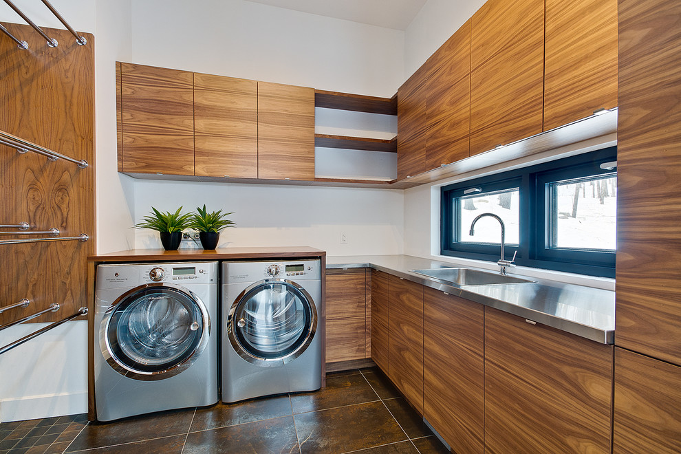 Moderne Waschküche in L-Form mit Einbauwaschbecken, flächenbündigen Schrankfronten, hellbraunen Holzschränken, Edelstahl-Arbeitsplatte, weißer Wandfarbe, Waschmaschine und Trockner nebeneinander, braunem Boden und grauer Arbeitsplatte in Montreal