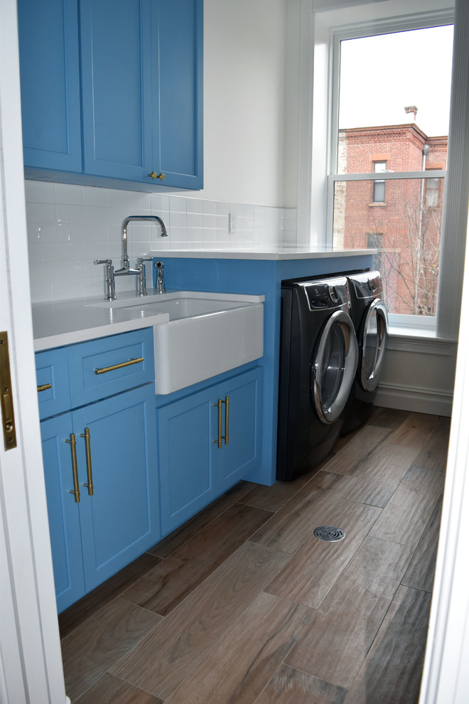 Inspiration for a mid-sized transitional ceramic tile and brown floor laundry room remodel in New York with a farmhouse sink, shaker cabinets, blue cabinets, quartz countertops, white walls, a side-by-side washer/dryer and white countertops