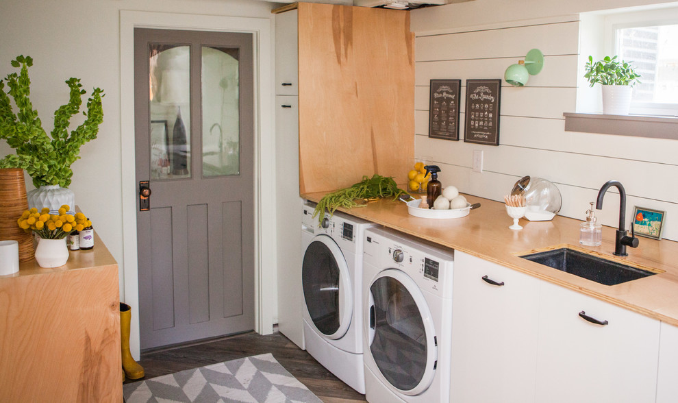 Photo of a medium sized farmhouse single-wall utility room in Denver with a submerged sink, flat-panel cabinets, white cabinets, wood worktops, white walls, dark hardwood flooring, a side by side washer and dryer and brown floors.