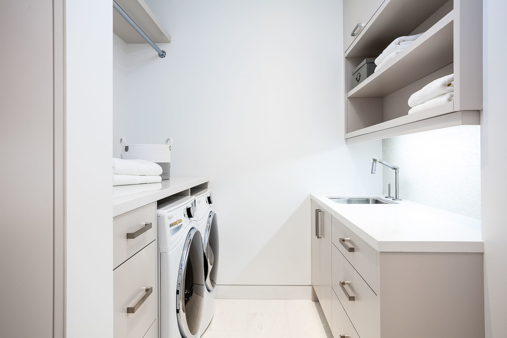 Modern inredning av en mellanstor parallell tvättstuga enbart för tvätt, med en undermonterad diskho, släta luckor, grå skåp, bänkskiva i kvarts, vita väggar, klinkergolv i porslin och en tvättmaskin och torktumlare bredvid varandra