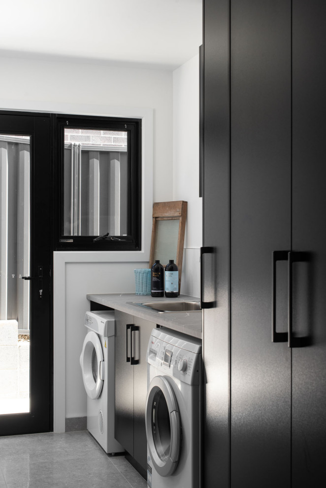 Moderne Waschküche mit flächenbündigen Schrankfronten, schwarzen Schränken, weißer Wandfarbe, Waschmaschine und Trockner nebeneinander, grauem Boden und grauer Arbeitsplatte in Hobart
