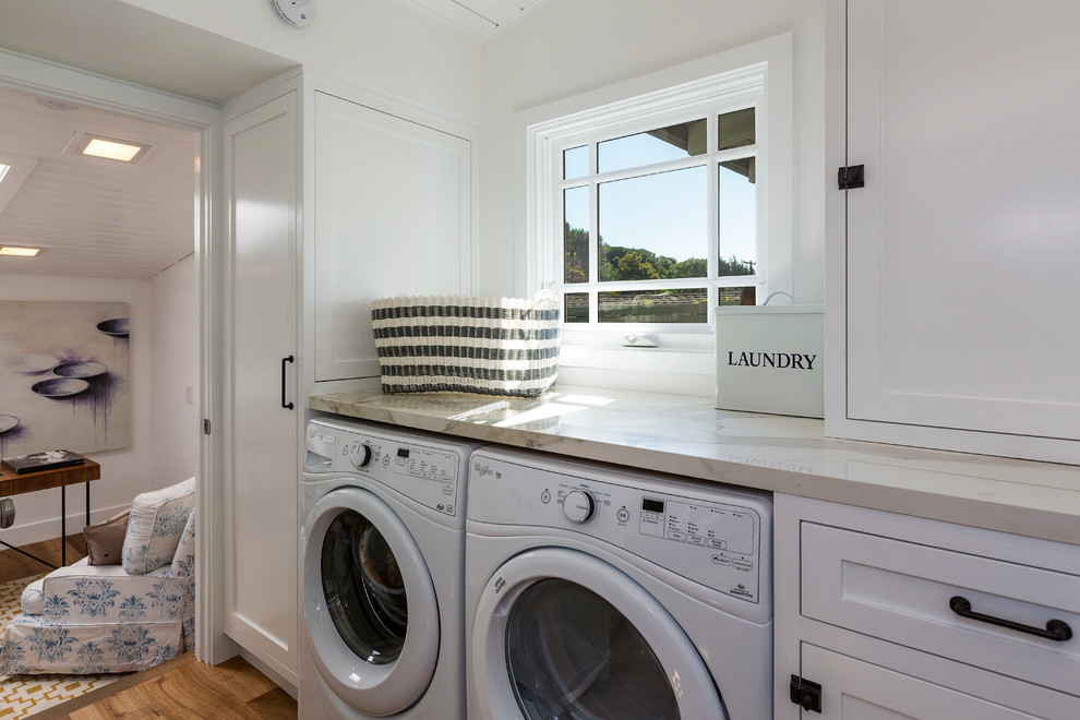 Foto de cuarto de lavado lineal costero de tamaño medio con armarios estilo shaker, puertas de armario blancas, paredes blancas, suelo de madera en tonos medios y lavadora y secadora juntas