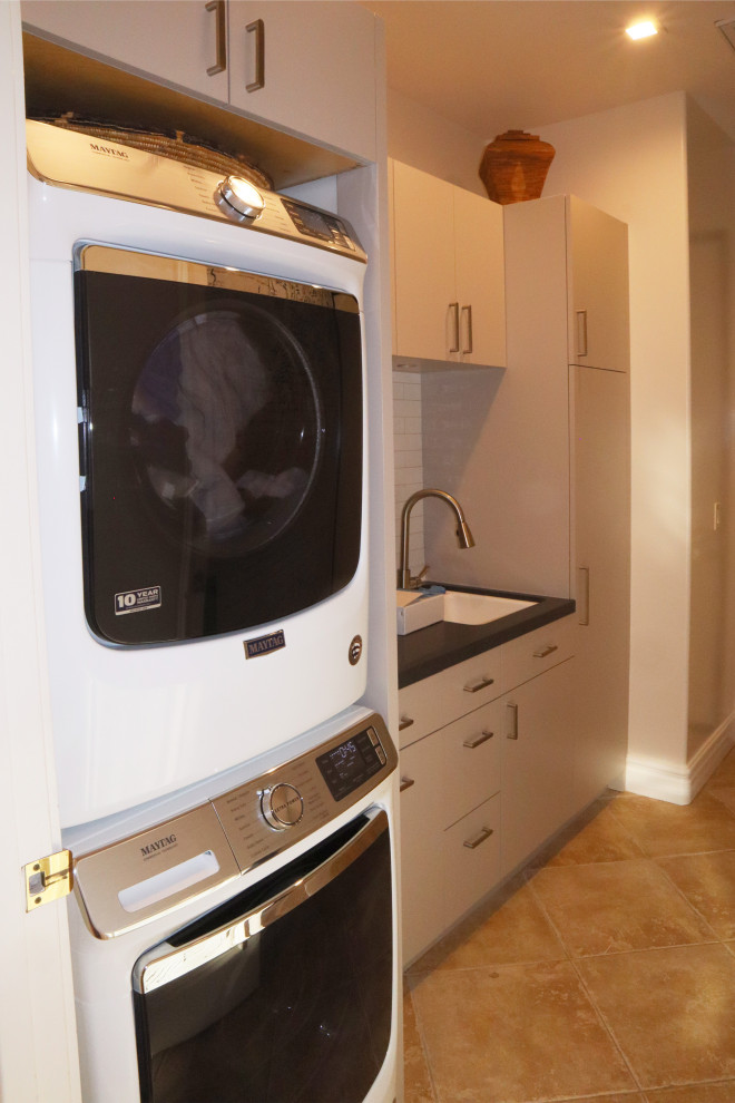 Example of a small trendy galley porcelain tile and beige floor dedicated laundry room design in Los Angeles with a drop-in sink, flat-panel cabinets, beige cabinets, quartz countertops, white walls, a stacked washer/dryer and black countertops