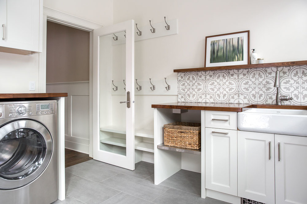 Dedicated laundry room - large traditional porcelain tile and gray floor dedicated laundry room idea in Other with a farmhouse sink, recessed-panel cabinets, white cabinets, wood countertops, white walls and a side-by-side washer/dryer