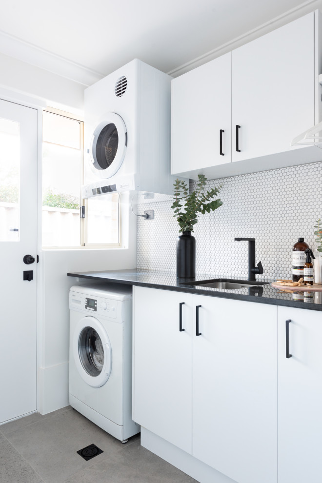 Inspiration for a medium sized contemporary single-wall utility room in Perth with a submerged sink, flat-panel cabinets, white cabinets, white splashback, white walls, porcelain flooring, a stacked washer and dryer, grey floors and black worktops.