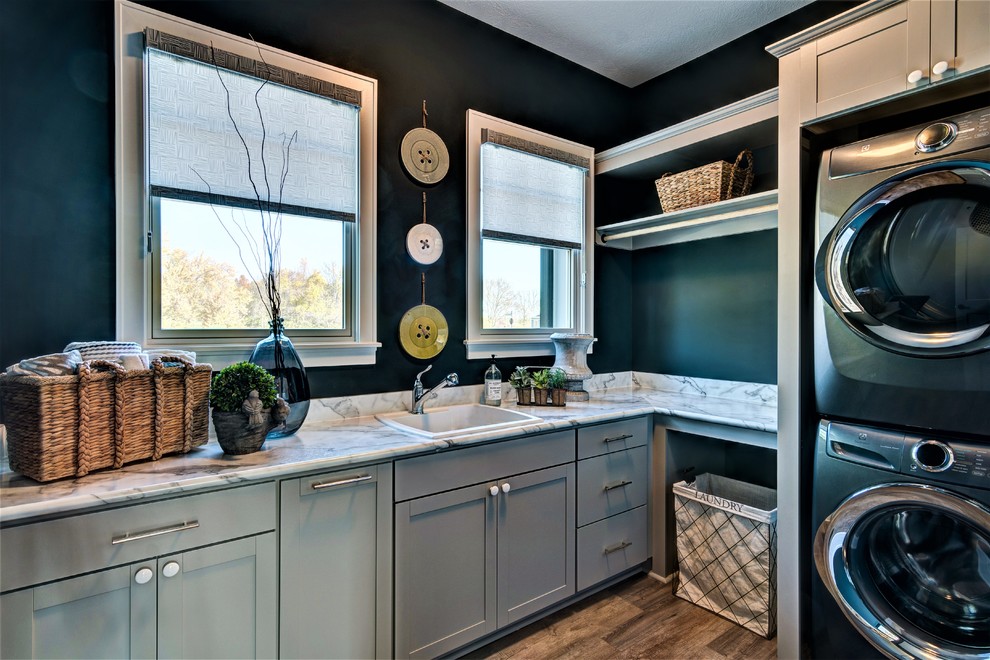 Inspiration for a transitional l-shaped medium tone wood floor and brown floor dedicated laundry room remodel in Indianapolis with a drop-in sink, shaker cabinets, gray cabinets, green walls and a stacked washer/dryer
