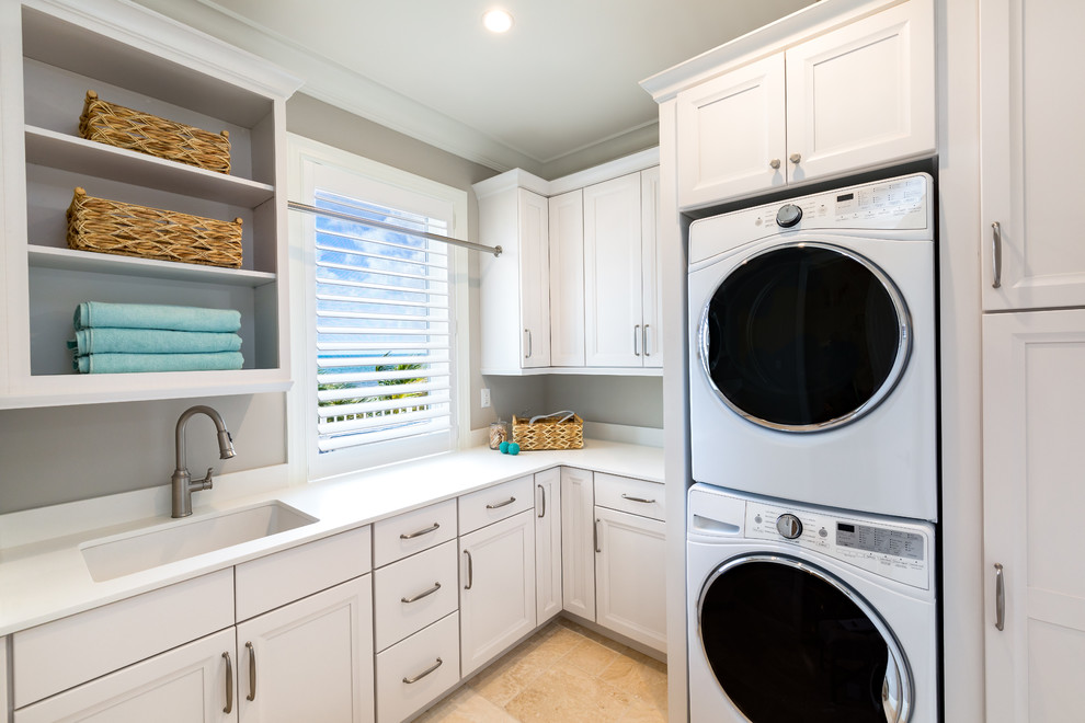 Design ideas for a beach style l-shaped separated utility room in Miami with a submerged sink, recessed-panel cabinets, white cabinets, grey walls, a stacked washer and dryer, beige floors and white worktops.