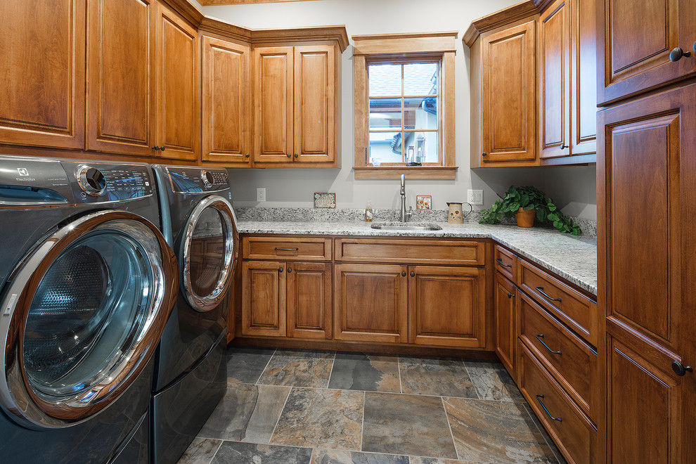 Rustik inredning av en mellanstor grå l-formad grått tvättstuga enbart för tvätt, med en undermonterad diskho, skåp i shakerstil, skåp i mellenmörkt trä, vita väggar, en tvättmaskin och torktumlare bredvid varandra och flerfärgat golv