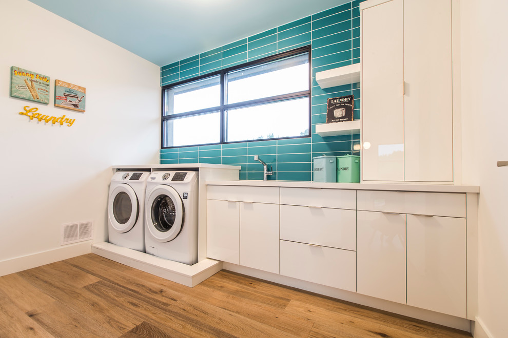 Bild på en funkis linjär tvättstuga, med släta luckor, vita skåp, vita väggar, mellanmörkt trägolv och en tvättmaskin och torktumlare bredvid varandra