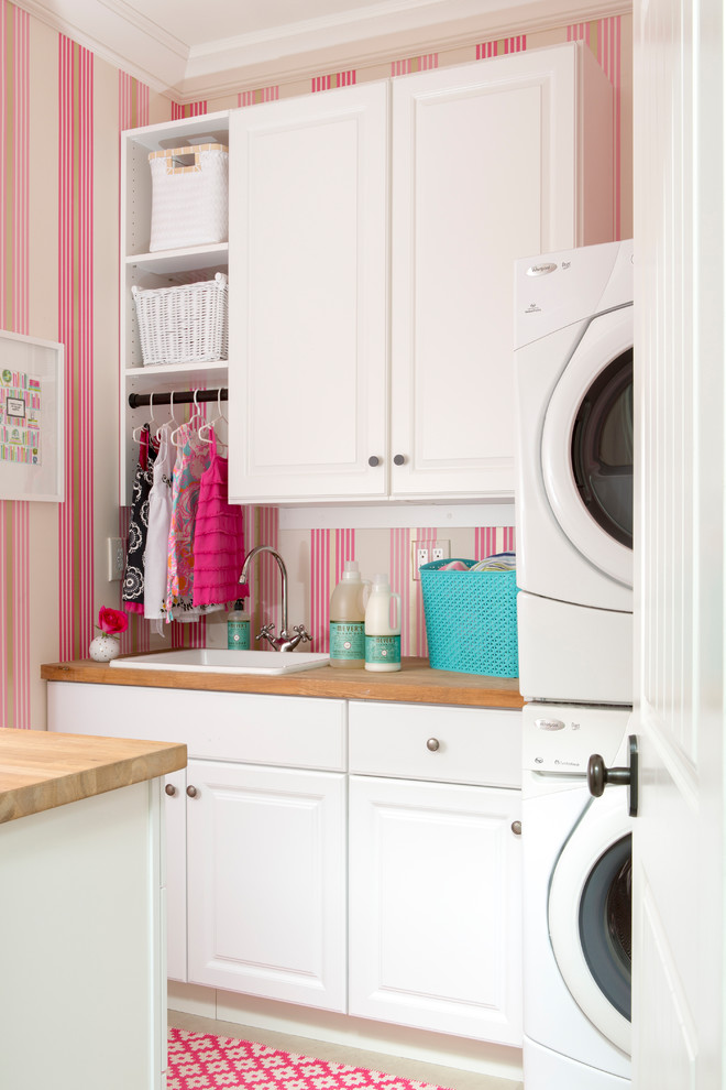 Zweizeilige Klassische Waschküche mit Einbauwaschbecken, profilierten Schrankfronten, weißen Schränken, Arbeitsplatte aus Holz, rosa Wandfarbe, Waschmaschine und Trockner gestapelt und rosa Boden in Los Angeles