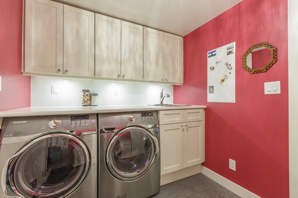 バンクーバーにある高級な中くらいなコンテンポラリースタイルのおしゃれな洗濯室 (ll型、ドロップインシンク、シェーカースタイル扉のキャビネット、白いキャビネット、ラミネートカウンター、ピンクの壁、コンクリートの床、左右配置の洗濯機・乾燥機) の写真