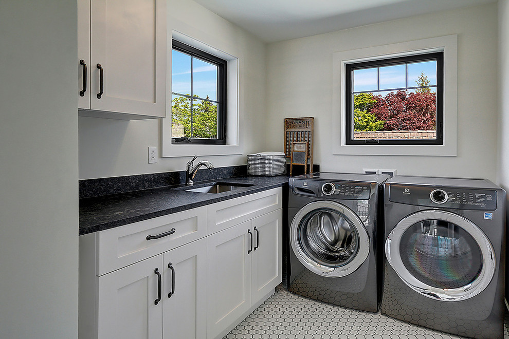 シアトルにあるトランジショナルスタイルのおしゃれな洗濯室 (L型、アンダーカウンターシンク、シェーカースタイル扉のキャビネット、白いキャビネット、白い壁、左右配置の洗濯機・乾燥機、白い床、黒いキッチンカウンター) の写真