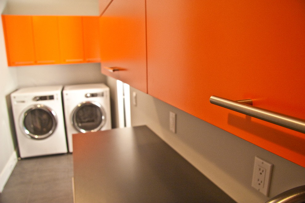 グランドラピッズにある小さなコンテンポラリースタイルのおしゃれな洗濯室 (I型、フラットパネル扉のキャビネット、オレンジのキャビネット、ラミネートカウンター、左右配置の洗濯機・乾燥機、黒いキッチンカウンター) の写真