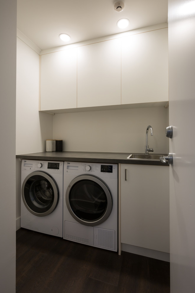 Modern inredning av en liten linjär tvättstuga enbart för tvätt, med en enkel diskho, släta luckor, vita skåp, laminatbänkskiva, vita väggar, laminatgolv, en tvättmaskin och torktumlare bredvid varandra och brunt golv