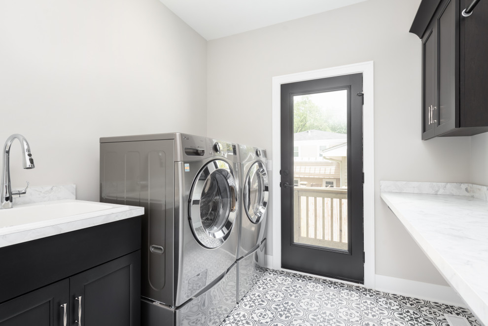 Inredning av en lantlig stor parallell tvättstuga enbart för tvätt, med en nedsänkt diskho, luckor med infälld panel, svarta skåp, grå väggar, klinkergolv i porslin, en tvättmaskin och torktumlare bredvid varandra och flerfärgat golv