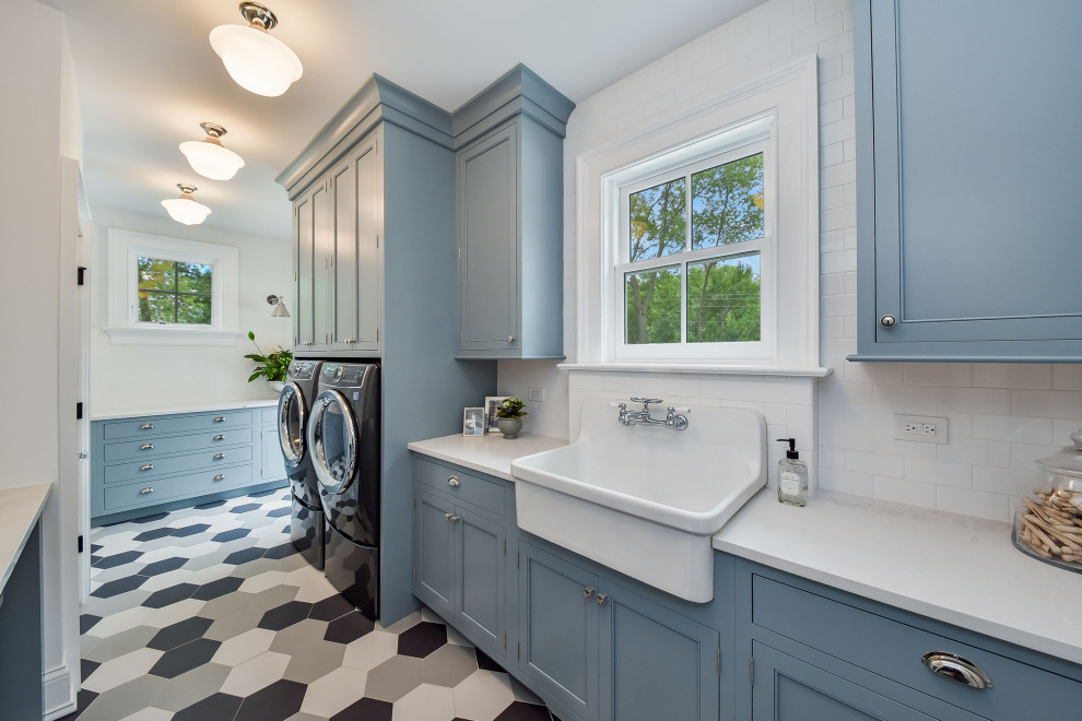 シカゴにある広いトランジショナルスタイルのおしゃれな洗濯室 (エプロンフロントシンク、フラットパネル扉のキャビネット、青いキャビネット、クオーツストーンカウンター、白いキッチンパネル、サブウェイタイルのキッチンパネル、白い壁、セラミックタイルの床、左右配置の洗濯機・乾燥機、青い床、白いキッチンカウンター) の写真