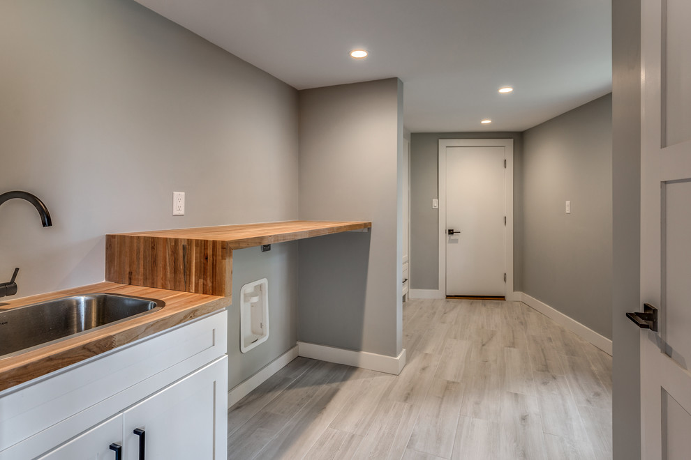 Example of a mid-sized trendy single-wall light wood floor and beige floor utility room design in Boston with a drop-in sink, shaker cabinets, white cabinets, wood countertops, gray walls and a side-by-side washer/dryer