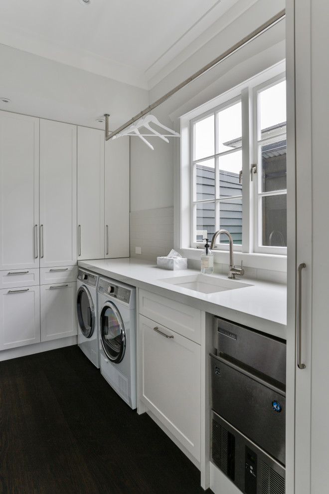 Foto på en vintage vita l-formad tvättstuga, med en undermonterad diskho, skåp i shakerstil, vita skåp, vitt stänkskydd, vita väggar, mörkt trägolv, en tvättmaskin och torktumlare bredvid varandra och brunt golv