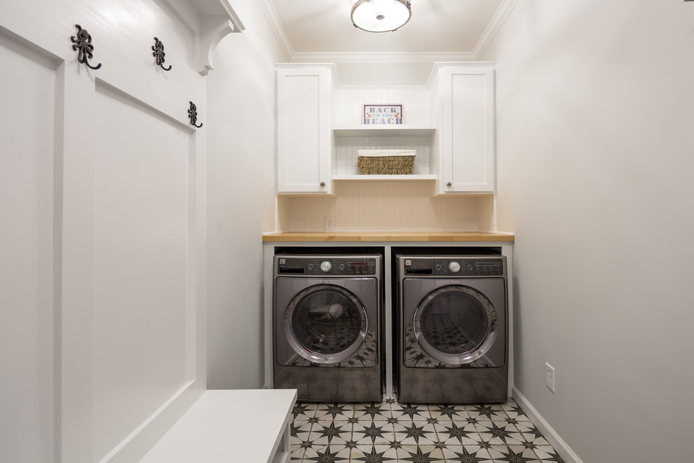 Klassisk inredning av en mellanstor bruna l-formad brunt tvättstuga enbart för tvätt, med skåp i shakerstil, vita skåp, träbänkskiva, grå väggar, klinkergolv i porslin, en tvättmaskin och torktumlare bredvid varandra och vitt golv