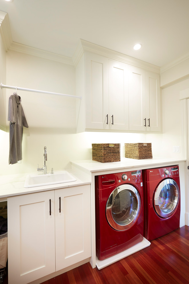 Immagine di una lavanderia multiuso stile americano di medie dimensioni con lavello da incasso, ante in stile shaker, ante bianche, top in quarzo composito, pareti bianche, parquet scuro, lavatrice e asciugatrice affiancate, pavimento rosso e top bianco