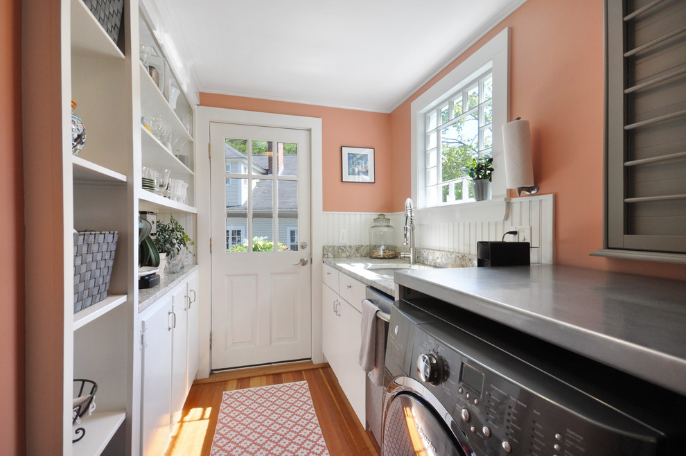 ボストンにあるお手頃価格の小さなトラディショナルスタイルのおしゃれな家事室 (ll型、アンダーカウンターシンク、フラットパネル扉のキャビネット、白いキャビネット、ステンレスカウンター、オレンジの壁、無垢フローリング、左右配置の洗濯機・乾燥機、茶色い床、マルチカラーのキッチンカウンター) の写真