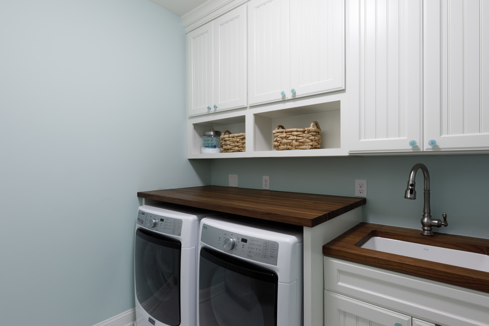 他の地域にある中くらいなトラディショナルスタイルのおしゃれな洗濯室 (白いキャビネット、青いキッチンパネル、ガラスタイルのキッチンパネル、淡色無垢フローリング、エプロンフロントシンク、木材カウンター、グレーの壁、左右配置の洗濯機・乾燥機、茶色いキッチンカウンター、落し込みパネル扉のキャビネット) の写真