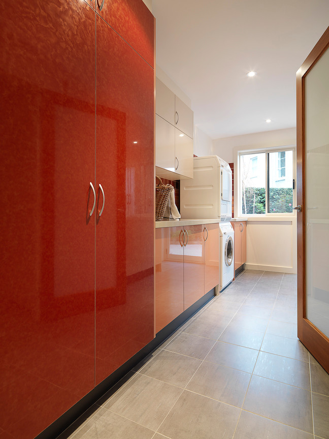 Photo of a large modern galley separated utility room in Melbourne with a submerged sink, flat-panel cabinets, engineered stone countertops, white walls, porcelain flooring, a stacked washer and dryer and red cabinets.