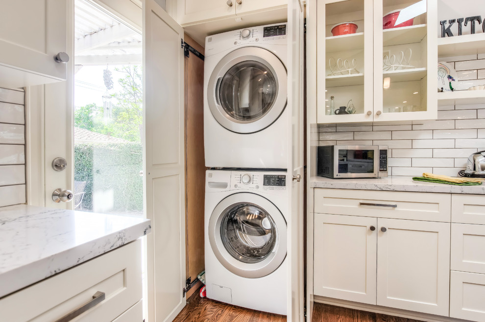 Inspiration for a transitional medium tone wood floor and brown floor laundry closet remodel in Los Angeles with a farmhouse sink, shaker cabinets, white cabinets, quartz countertops, white walls, a stacked washer/dryer and white countertops