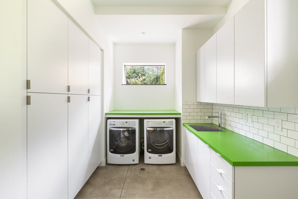 Moderne Waschküche in U-Form mit grüner Arbeitsplatte, flächenbündigen Schrankfronten, weißen Schränken, weißer Wandfarbe, Betonboden, Waschmaschine und Trockner nebeneinander, grauem Boden, Unterbauwaschbecken und Laminat-Arbeitsplatte in Denver